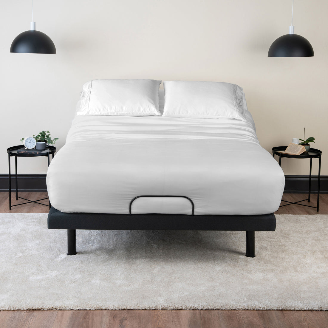 A mattress made up with white sheets and pillows on a dark adjustable bed base, between two black side tables under pendant lights