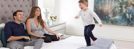 Family relaxing together on a bed in a bright bedroom while a child jumps beside them