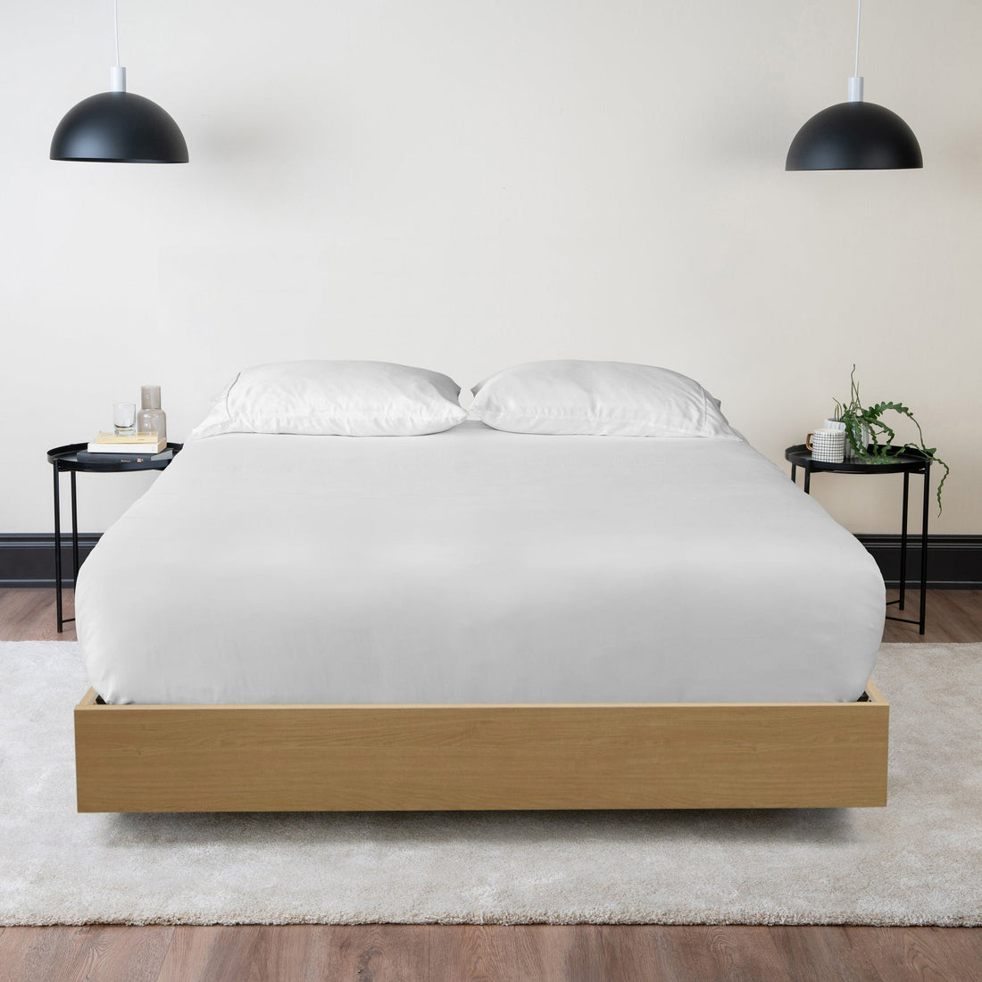 A made bed with white sheets and two pillows on a light wood platform frame, between two black side tables
