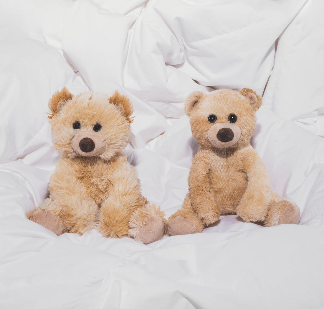 Two teddy bears sitting side by side on a rumpled white duvet