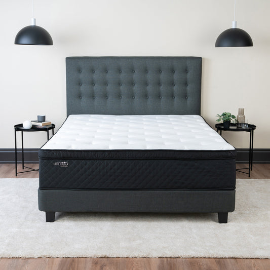 Hamuq Original Hybrid mattress with a white quilted top and black side panel on a dark grey upholstered bed with a tufted headboard, between two black side tables