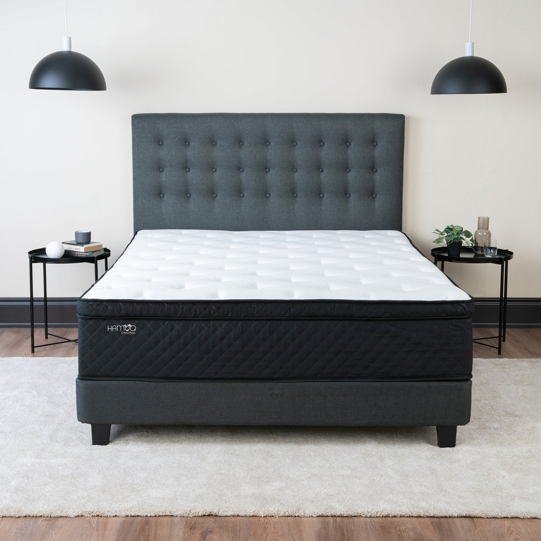 Hamuq Original Hybrid mattress with a white quilted top and black side panel on a grey tufted upholstered bed frame, between two black side tables